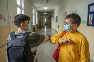 
		Two schoolchildren greet each other at school with masks and elbows.
	